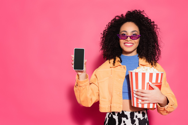 uśmiechnięta Afroamerykanka w okularach przeciwsłonecznych trzymająca wiadro popcornu i smartfona z czystym ekranem na różowym  - Zdjęcie, obraz