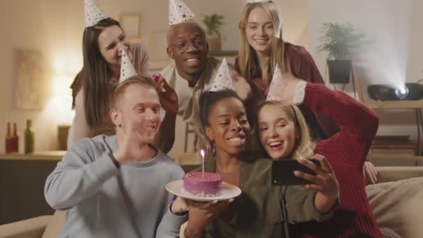 Średnie ujęcie szczęśliwej grupy ludzi w kapeluszach imprezowych trzymających mały tort urodzinowy ze świecą i machających rękami podczas robienia selfie lub filmowania siebie na telefonie komórkowym - Materiał filmowy, wideo