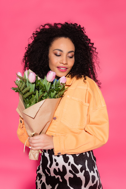 curly african american woman holding bouquet of flowers isolated on pink  - Zdjęcie, obraz