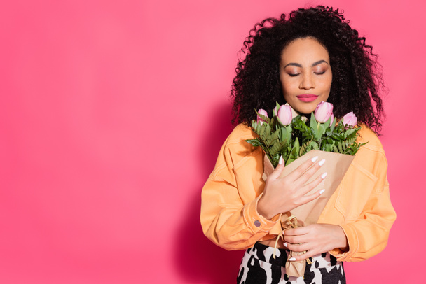 african american woman with closed eyes smelling flowers on pink - Foto, imagen