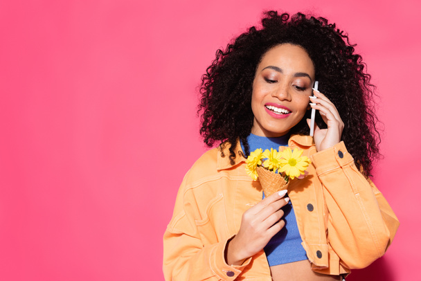 happy african american woman talking on smartphone and holding waffle cone with flowers on pink  - Zdjęcie, obraz
