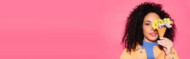 curly african american woman covering eye while holding holding waffle cone with flowers on pink, banner - Foto, afbeelding