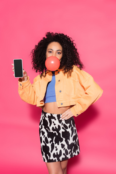 кучерява афроамериканська жінка дме бульбашкову гумку і тримає смартфон з порожнім екраном на рожевому
  - Фото, зображення