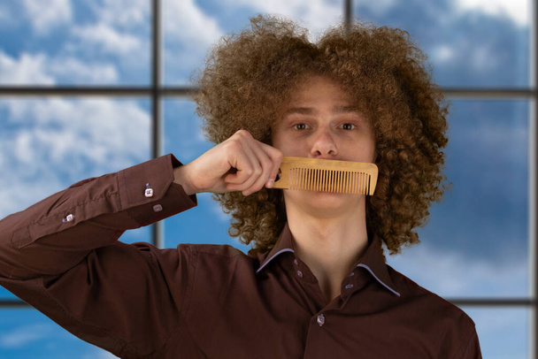 A long-haired curly-haired guy in a brown shirtuses a wooden comb. Emotions before a haircut in a hairdresser. - Foto, immagini