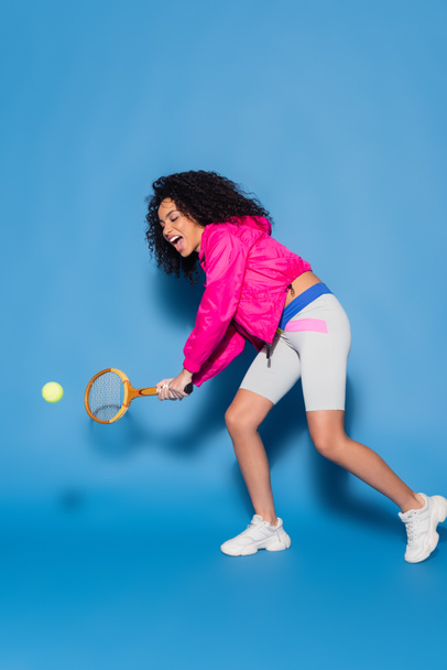 full length of excited african american woman playing tennis on blue - Foto, Imagem