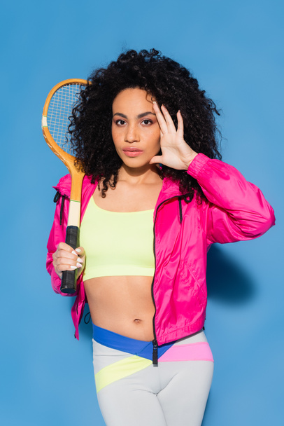 curly african american woman in crop top posing with tennis racket and looking at camera on blue - Foto, imagen