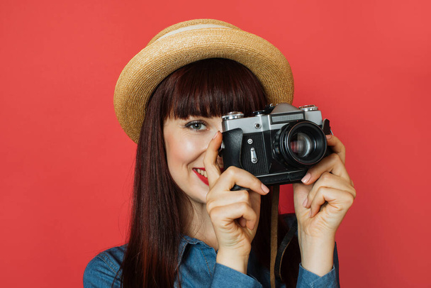 Close up of beautiful young woman photographer in straw hat taking a picture and looking in viewfinder on retro digital mirrorless photo camera, posing on studio isolated red background - 写真・画像