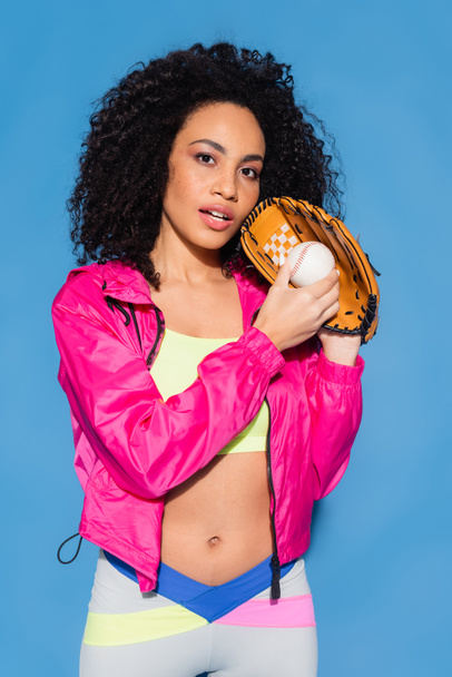 curly african american woman in leather glove holding baseball on blue - 写真・画像