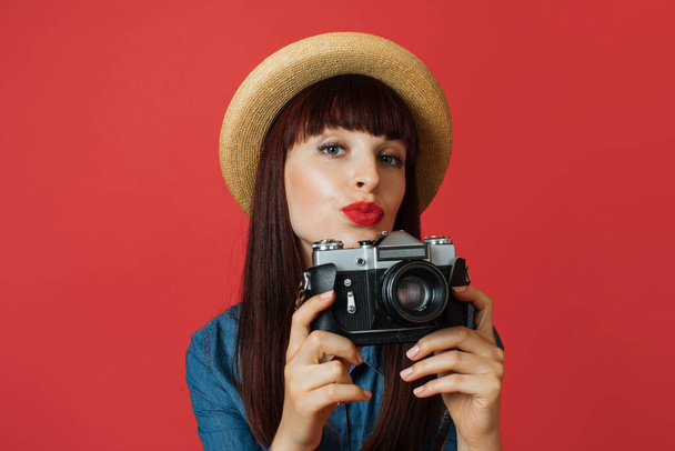 Hobby, taking photos, travel concept. Gorgeous young woman in hat, photographer amateur, smiling and making air kiss, taking picture on old vintage camera, on colorful red studio background. - Foto, Imagem