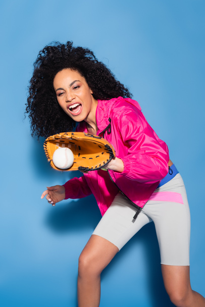 excited african american woman in leather glove catching baseball on blue - Fotografie, Obrázek