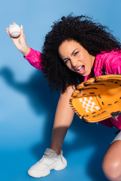 ενθουσιασμένοι αφροαμερικανός γυναίκα σε δερμάτινο γάντι κρατώντας μπέιζμπολ στο μπλε - Φωτογραφία, εικόνα