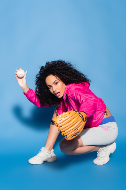 curly african american woman in leather glove throwing baseball on blue - Φωτογραφία, εικόνα