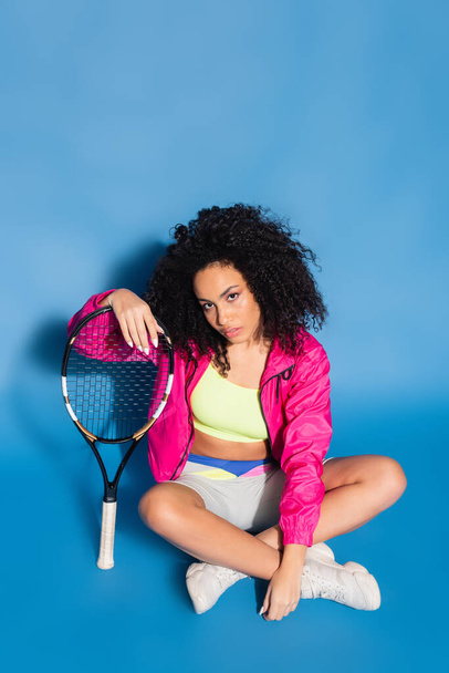 afro-américaine assise près de raquette de tennis sur bleu - Photo, image