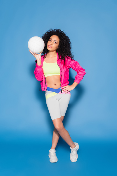 full length of sporty african american woman in pink jacket holding volleyball while posing with hand on hip on blue - Foto, Bild