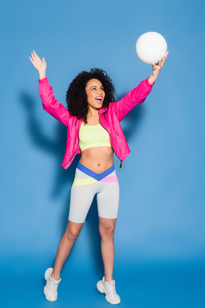 full length of amazed african american woman in pink jacket and crop top playing volleyball on blue - Фото, изображение