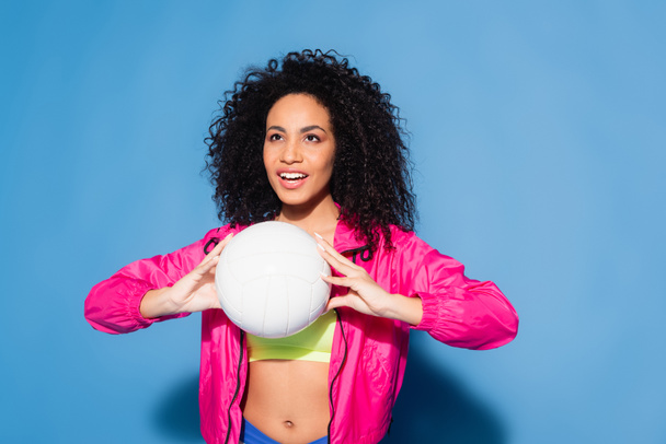 happy african american woman in pink jacket and crop top playing volleyball on blue - Zdjęcie, obraz