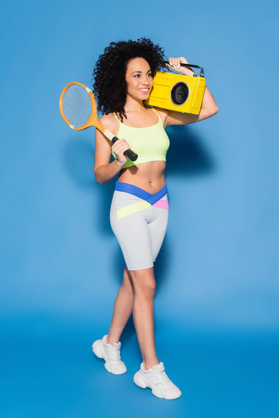 full length of happy african american woman holding boombox and tennis racket on blue - Valokuva, kuva
