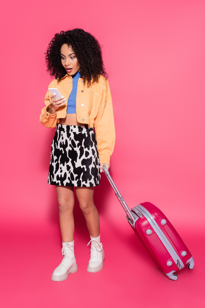 full length of shocked african american woman using smartphone near suitcase on pink  - Foto, Imagen