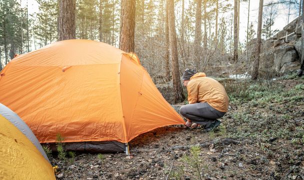 オスハイカーは森の中に明るいオレンジ色のテントを張っています。観光、ハイキング、自然の中での滞在の概念. - 写真・画像
