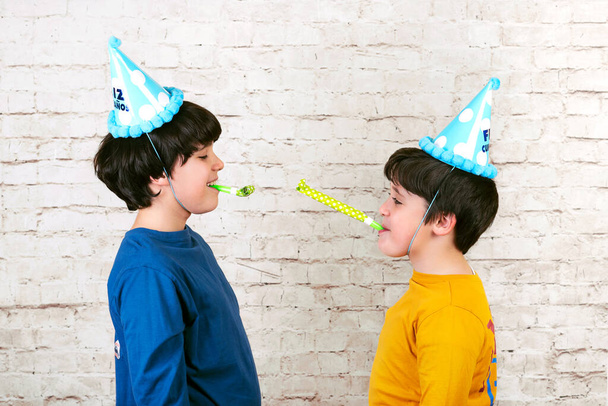 divertenti bambini con cappello da festa che soffia in un corno di partito su sfondo muro di mattoni bianchi - Foto, immagini