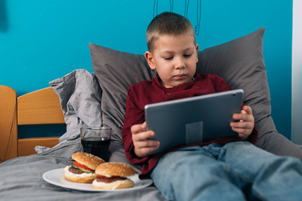 young boy eating unhealthy food and using digital tablet while sitting in his room - Foto, Bild