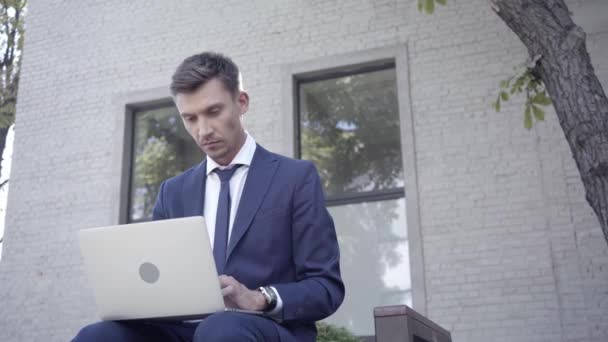 Biznesmen korzystający z laptopa i siedzący na ławce na zewnątrz  - Materiał filmowy, wideo