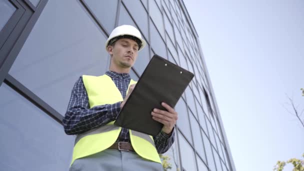 Builder in hard hat writing on clipboard outdoors  - Footage, Video