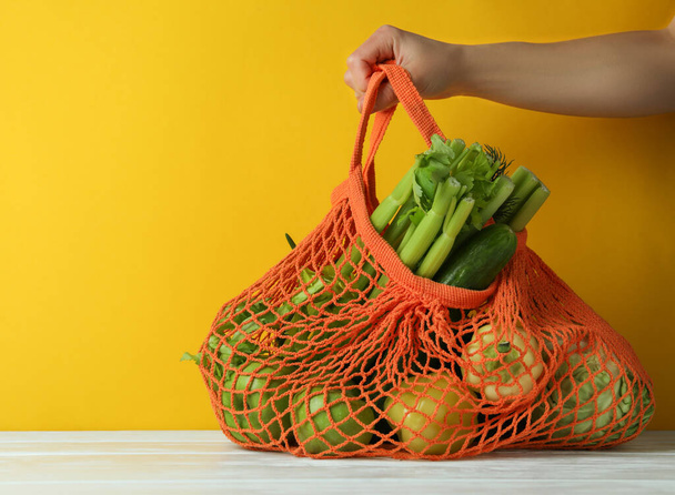 Female hand hold string bag with green food on yellow background - 写真・画像