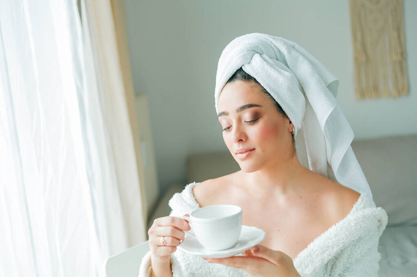 シャワーの後、頭にタオルをつけたバスローブの若い女性がコーヒーを飲み、窓から外を眺めます。朝だ。朝食. - 写真・画像