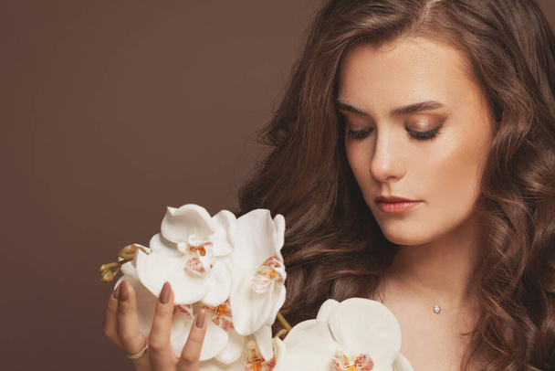 Gesunde junge Model-Frau mit perfekter Haut, braunem lockigem Haar und weißen Orchideenblüten. Gesichtsbehandlung und Hautpflegekonzept - Foto, Bild