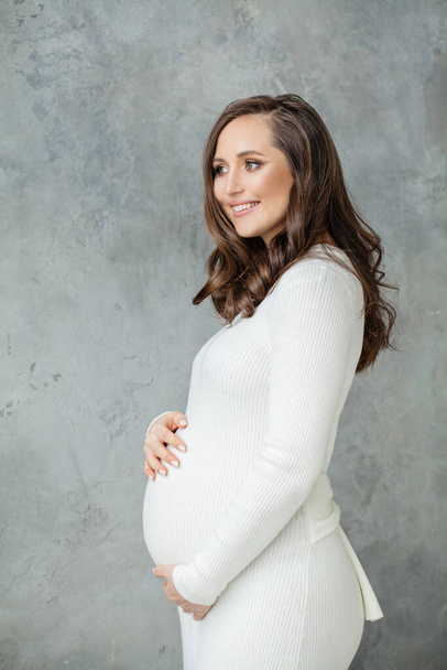 Красивая улыбающаяся беременная женщина стоит на сером фоне - Фото, изображение