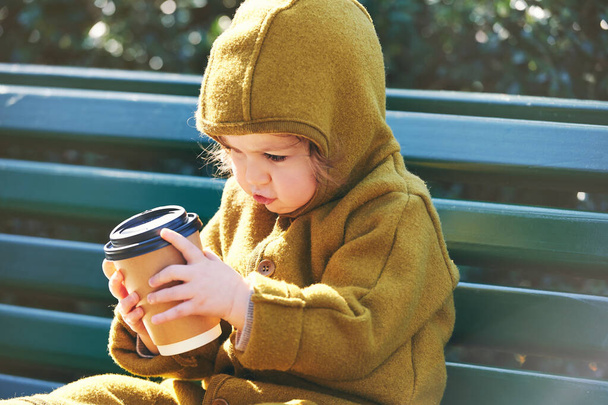 Divertido niño pequeño descansando en el banco, sosteniendo una taza de papel de café para llevar - Foto, Imagen