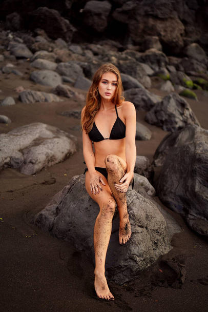 Bikini girl relaxing on the rocky black beach. Young beautiful woman in fashionable swimsuit. Summer vibes - Foto, Bild
