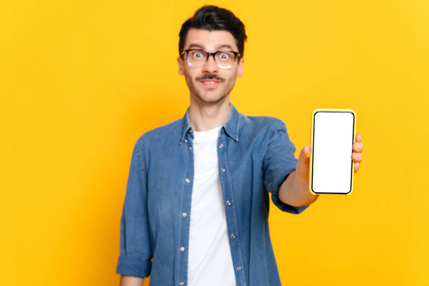 Defocused caucasian smiling guy with glasses in denim shirt, looks at the camera, holds smartphone with white empty screen for your presentation in hand, copy space, isolated orange background - Φωτογραφία, εικόνα