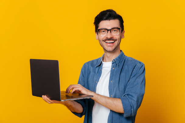 Atrakcyjny, zadowolony, stylowy, nieogolony, inteligentny biały facet w okularach z otwartym laptopem w ręku, uśmiechnięty do aparatu, stojący naprzeciw odizolowanej pomarańczy - Zdjęcie, obraz