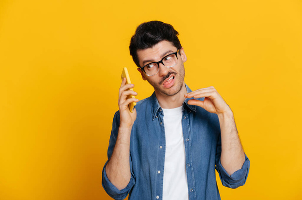 Phone call by cellphone. Attractive charismatic stylish guy has an unpleasant phone conversation, ignores the interlocutor, makes blah blah gesture, stands on isolated orange background, looks away - Photo, Image