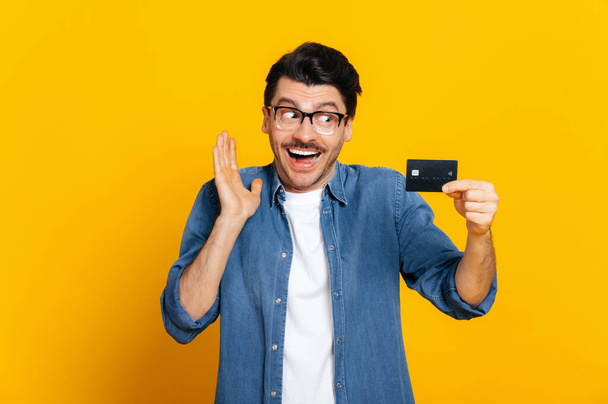 Blij opgewonden Kaukasische ongeschoren knappe stijlvolle man met bril, houdt credit card in de hand, kijkt haar verbaasd aan, staat op een geïsoleerde oranje achtergrond - Foto, afbeelding