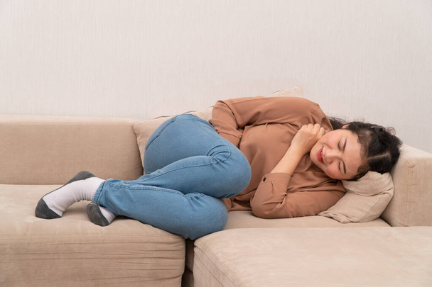 Unzufriedene Asiatin, die auf dem Sofa sitzt und sich an Bauchschmerzen festhält. Bauchschmerzen, die von Menstruation, Durchfall oder Verdauungsstörungen herrühren. Krankheits- und Gesundheitskonzept - Foto, Bild