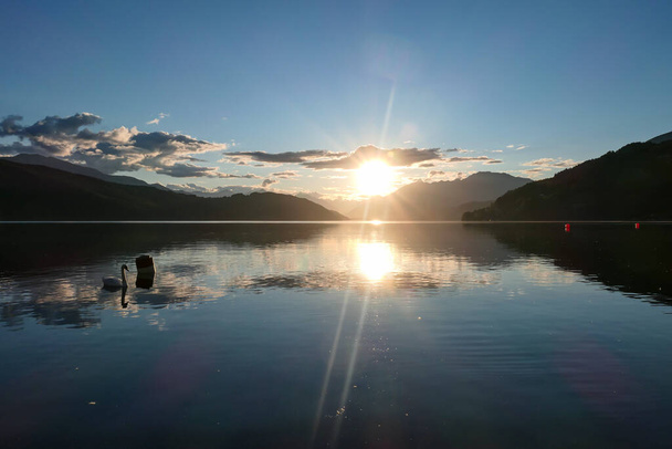Egy hattyú kelt át a Millstaetter tavon Ausztriában naplementekor. A tavat magas Alpok veszi körül. A tó nyugodt felszíne visszaveri a napsugarakat. A nap a sűrű felhők mögé bújik. Nyugodj meg. - Fotó, kép
