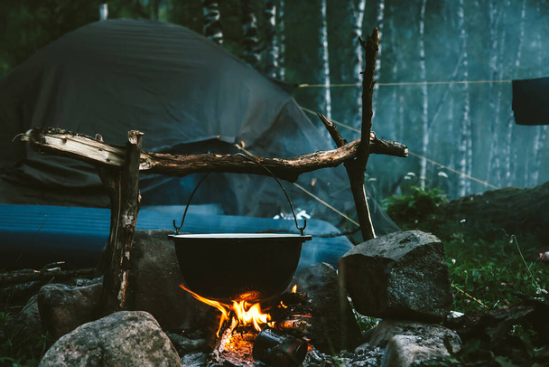 Kettle on fire near tent in forest at night. Beautiful campfire in tourist camp into wild. Survival in taiga. Cauldron above bonfire. Smoke from fire among trees. Cooking over campfire. - Valokuva, kuva