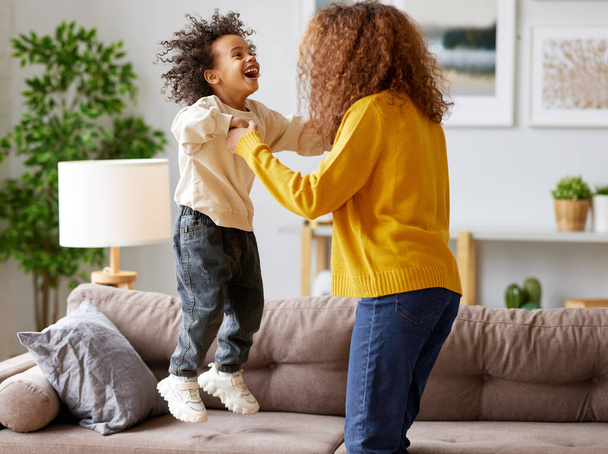 Crazy weekend with mom. Joyful afro american family mother and little son holding hands and jumping on sofa in living room at home. Mom and child in casual wear playing together - Foto, Imagem