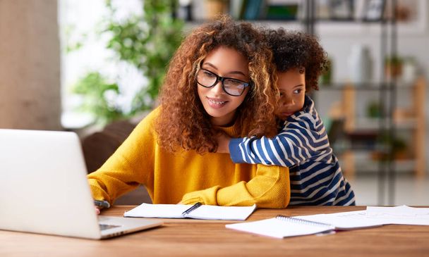 Arbeit mit Kindern. Junge fokussierte Mutter trägt eine Brille mit Laptop und denkt über ihre Arbeitsaufgabe nach, während ihr kleiner Sohn sie sanft umarmt. Kinderbetreuungskonzept - Foto, Bild