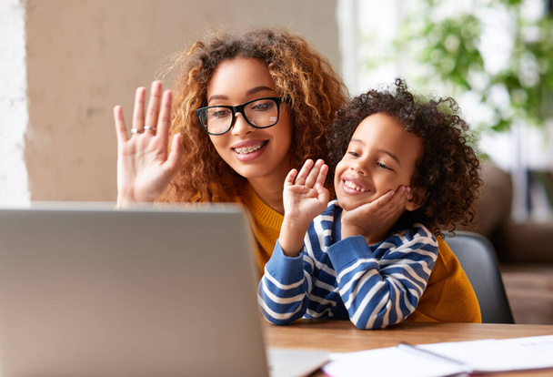 Onnellinen afro amerikkalainen perhe, nuori positiivinen äiti ja söpö poika, jolla on videopuhelu sukulaisten, isän tai isovanhempien kanssa, heiluttaen webbikameralla kannettavalla tietokoneella ja hymyillen, kun hän istuu pöydän ääressä kotona - Valokuva, kuva
