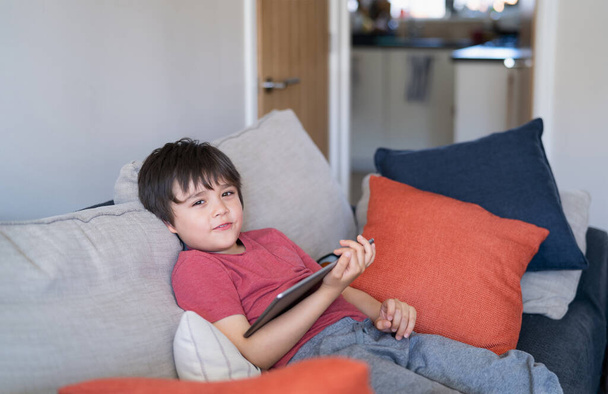 Authentic Kid sitting on sofa watching cartoons or playing games on tablet,Child boy using digital pad learning lesson on internet,Home schooling,Distance learning online education concept - Photo, Image