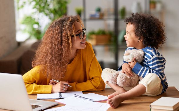 Cute little african boy with curly hair sitting on table, holding teddy bear and talking with smiling mom while she working remotely on laptop from home, selective focus on people. Childcare concept - Photo, Image