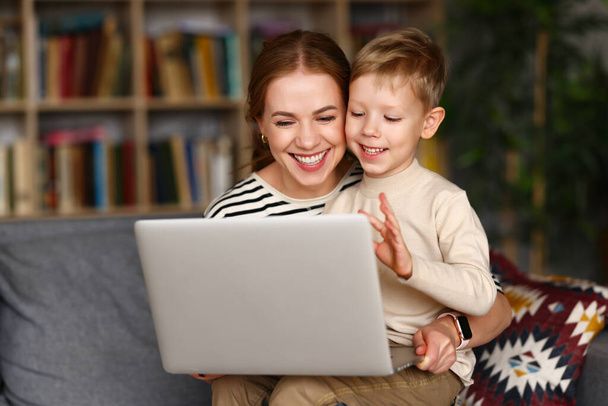 Happy family young loving mother and cute little boy son looking at computer screen, smiling and waving while having video call with family on laptop, relaxing together on sofa in living room at home - 写真・画像