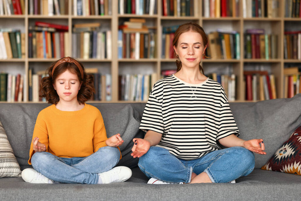 Junge Mutter und Tochter in Freizeitkleidung meditieren mit geschlossenen Augen, während sie im Lotus sitzen und gemeinsam auf dem Sofa im Wohnzimmer posieren. Ruhige schöne Familie praktiziert Yoga während der Quarantäne - Foto, Bild