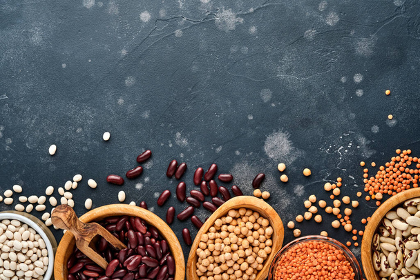 Legumes, red lentils, chickpea, red and white beans assortment in different bowls on dark black table. Diet for weight loss and fight against cholesterol concept. Top view with copy space. - Foto, Imagem