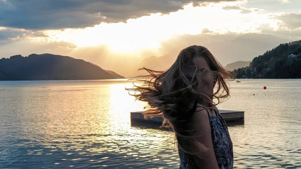 A woman in a blue dress flips her long, brown hair by the shore of Millstaettersee lake. The sun is setting behind high Alps. Calm surface of the lake reflects the orange sky and the mountains. Fun - Photo, Image