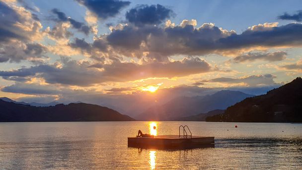 Een man op een houten platform drijvend op het MIllstaetter meer tijdens de zonsondergang. De zon gaat onder achter de hoge Alpen. Kalme oppervlakte van het meer weerspiegelt de oranje lucht. Mediterende ale kalmerende. - Foto, afbeelding